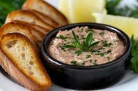 Creamy salmon pate served in dark ceramic bowl with toasted baguette slices, fresh herbs and lemon wedges on white plate, close up view of appetizer spread.