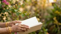 Senior woman with South Asian ethnicity reading an open book outdoors in a garden with blooming flowers, suitable for education, literacy, and wellness content.