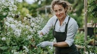 Mature Caucasian female gardener in apron and gloves tending to white flowering shrubs in lush garden setting, expressing joy while pruning and maintaining plants.