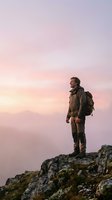 Male hiker with backpack standing on rocky mountain peak at sunrise, gazing at misty valley landscape below. Adventure travel and outdoor exploration concept.