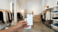Person holding blank white price tag or label in modern clothing retail store with racks of apparel and neutral interior design for marketing and branding purposes.