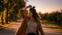 Young woman with long brown hair shielding eyes from golden sunset light while standing on rural road surrounded by trees during warm evening hour.