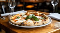 Fresh Neapolitan margherita pizza with melted mozzarella, tomato sauce and basil leaves served on white plate at rustic wooden restaurant table with wine glasses.