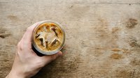 Caucasian hand holding iced coffee drink with caramel swirl on rustic wooden surface, overhead view for beverage and lifestyle content.