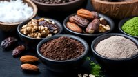 Healthy superfood ingredients in black ceramic bowls on dark background - raw cacao powder, matcha tea, nuts, dates and natural sweeteners.
