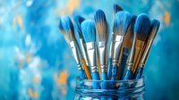 Paint brushes with blue bristles arranged in glass jar against turquoise background with soft bokeh effect, artistic supplies for creative work and painting.