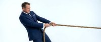 Caucasian male professional in dark navy business suit pulling rope with determined expression, symbolizing business challenge, competition, and corporate strength against white background.