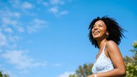 Young woman with dark curly hair and medium skin tone wearing light sleeveless top, smiling outdoors against clear blue sky with clouds and tropical foliage, expressing joy and happiness.