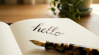 Fountain pen resting on open notebook with handwritten hello in elegant calligraphy script, warm wooden desk background with green plant.