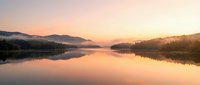 Serene lake at sunrise with misty mountains and forested shoreline reflected in calm water, peaceful nature landscape for meditation and wellness projects.