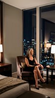 Elegant blonde businesswoman in black dress relaxing in luxury hotel room with city skyline view at night for corporate travel and hospitality concepts.
