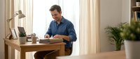 Middle-aged Caucasian man in blue shirt working at wooden desk in bright home office with plants and natural lighting for remote work concepts.