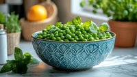 Fresh green peas in decorative turquoise ceramic bowl with mint leaves on marble counter, natural light setting with herbs and citrus in background.
