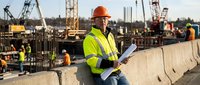 Construction worker in safety gear reviewing blueprints at busy building site with cranes and equipment in background during daytime operations.