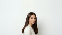 Young woman with long dark brown hair and fair complexion, wearing a light beige or cream-colored shirt, posed against a neutral white background, suitable for portrait and professional use.