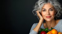Confident mature Caucasian woman with natural gray curly hair smiling while holding fresh citrus fruits, wearing casual gray top against dark background.