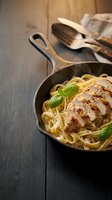 Grilled chicken breast with fettuccine pasta in cast iron skillet garnished with fresh basil leaves on rustic wooden table for restaurant menu design.