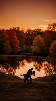 Romantic couple silhouette dancing by lake during golden autumn sunset with colorful fall foliage reflected in calm water for wedding and love concepts.