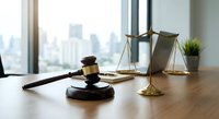 Wooden gavel and brass scales of justice on desk in modern law office with city skyline view. Legal consultation and courtroom proceedings concept.