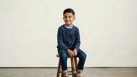Young boy with dark hair and medium skin tone, wearing casual denim clothing, sitting on wooden stool against white background. Suitable for family, children, lifestyle, and educational content.