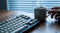 Caucasian hand holding a steaming coffee mug beside a keyboard on a wooden desk near window blinds, morning work routine concept.