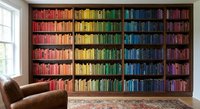 Rainbow colored books arranged by hue on floor-to-ceiling bookshelf in modern home library with leather armchair and window creating organized literary display.