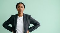 Young woman of African descent wearing a dark blazer over a white shirt, standing with hands on hips against a mint green background, suitable for corporate, business, and professional website design.