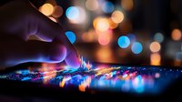 Finger touching digital screen with colorful light visualization against blurred bokeh background, creating interactive technology experience.