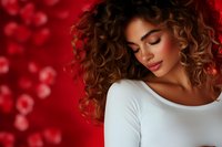 Young woman with curly brown hair and natural makeup against red bokeh background, wearing white top, eyes closed in serene expression. Suitable for beauty and fashion.