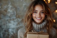 Young mixed race woman with bright smile holding wrapped gift box, wearing cozy knit sweater against bokeh lights background. Holiday season portrait.