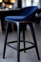 Modern bar stool with curved blue leather seat and black wooden base against blurred restaurant background, contemporary minimalist furniture design.