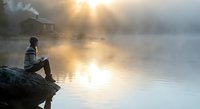 Young man sitting on rocky shore reading book by misty lake at sunrise with cabin in background creating peaceful morning atmosphere.