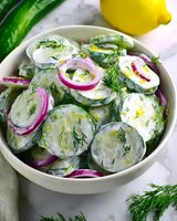 Fresh cucumber salad with creamy dressing, red onion rings and dill in white ceramic bowl. Bright summer appetizer with lemon garnish on marble surface.