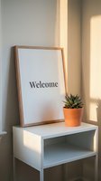 Wooden framed welcome sign leaning on a white shelf beside a small succulent in a terracotta pot, bathed in warm natural sunlight.