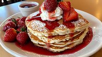 Stack of fluffy pancakes topped with fresh strawberries, whipped cream, powdered sugar, and strawberry sauce on white plate, served with extra syrup on side.