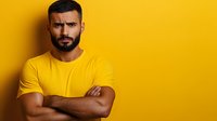 Middle Eastern man with beard wearing yellow t-shirt stands with crossed arms against vibrant yellow wall, expressing serious skeptical emotion. Perfect for business or lifestyle content.