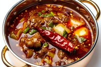 Spicy Asian hot pot with beef chunks, chili peppers, and vegetables in rich red broth garnished with fresh herbs and sesame seeds served in traditional metal pot.