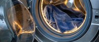 Open washing machine drum with blue denim jeans inside, modern laundry appliance with stainless steel interior and warm lighting for household cleaning concepts.
