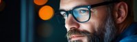 Bearded man in glasses illuminated by blue light from screen with orange bokeh background, side view portrait showing focused concentration.