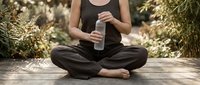 Woman in black athletic wear sitting cross-legged on wooden deck holding water bottle, surrounded by green plants and natural sunlight, wellness and mindfulness lifestyle.