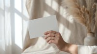 Woman holding blank white card mockup in natural sunlight with pampas grass decor for branding design templates and stationery presentation concepts.
