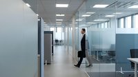 Young professional man in business suit walking through modern office corridor with glass partitions and contemporary lighting.
