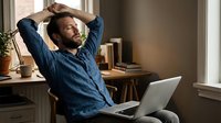 Relaxed Caucasian man stretching at home office desk with laptop, taking a break from remote work, eyes closed, casual blue shirt.