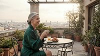 Mature woman with gray hair and light skin tone, wearing a green jacket, enjoying coffee and pastry on a rooftop terrace with cityscape views. Lifestyle moment capturing leisure and relaxation.