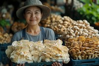 Asian female farmer in hat holding fresh organic oyster mushrooms at local market, surrounded by variety of cultivated fungi displayed for sale.