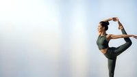 Young Asian woman practicing yoga dancer pose outdoors against cloudy sky wearing green athletic wear for fitness wellness and mindfulness training.