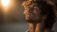 Young African American man with curly hair basking in warm golden sunlight, eyes closed in peaceful meditation or reflection.
