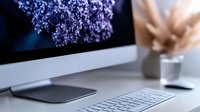 Minimalist workspace with computer monitor, wireless keyboard and mouse on white desk, decorated with lilac flowers in soft morning light.