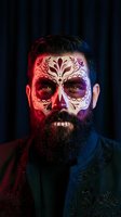 Bearded man with ornate sugar skull face paint featuring floral patterns in dramatic red and blue lighting for Day of the Dead celebration.