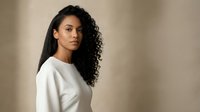 Young African American woman with curly hair wearing white sweater looking over shoulder against neutral beige background for lifestyle and fashion content.